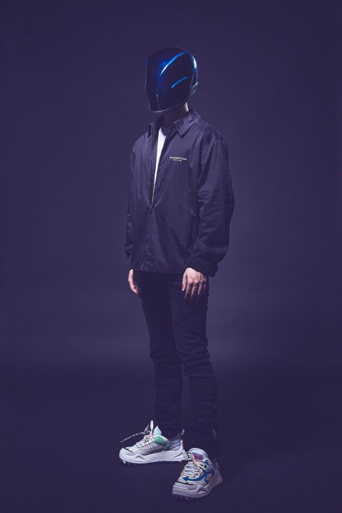 Full-length studio portrait of a person wearing a glossy blue helmet and a black coach jacket over dark trousers and sneakers.