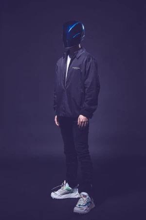 Full-length studio portrait of a person wearing a glossy blue helmet and a black coach jacket over dark trousers and sneakers.