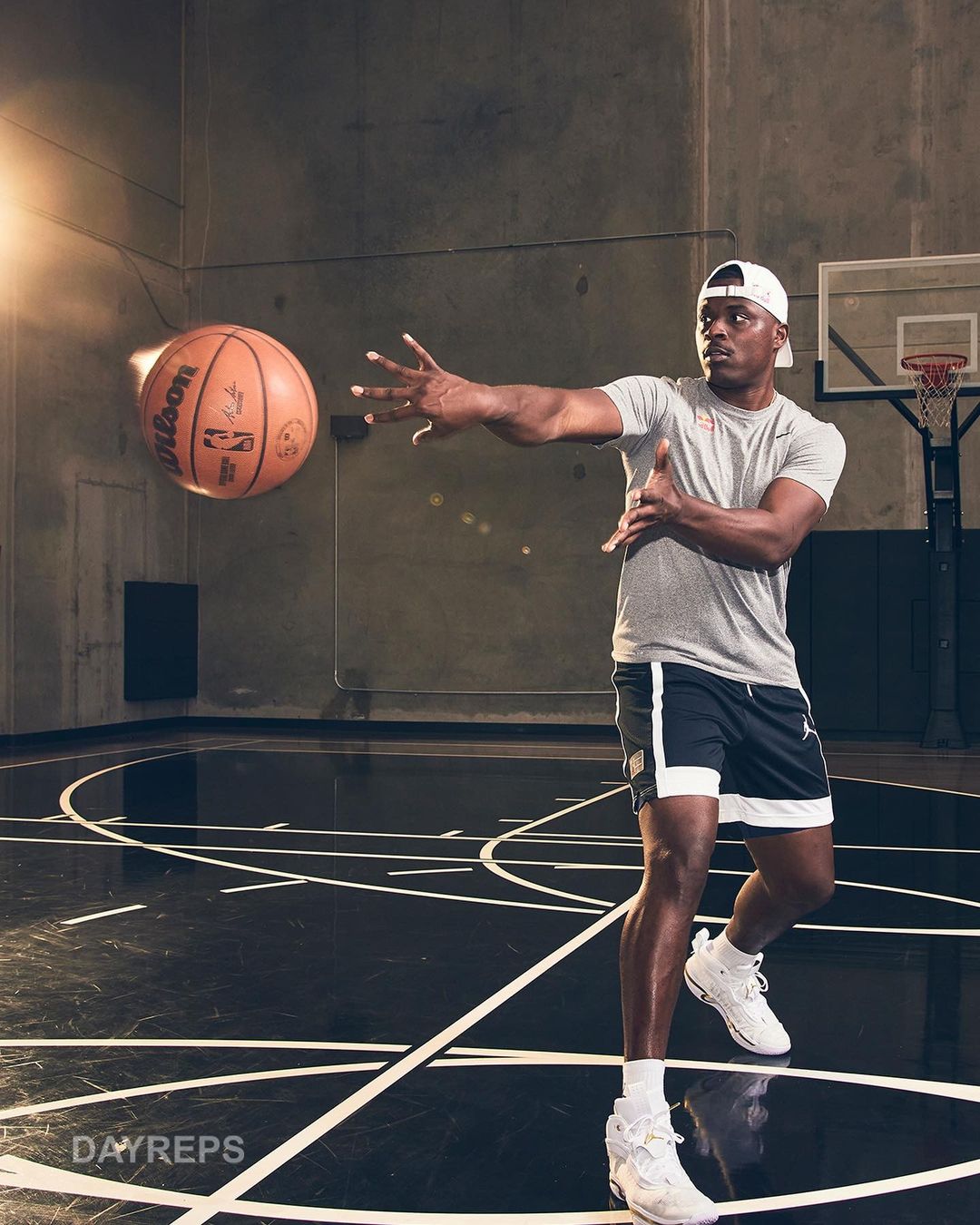 An African American man on a basketball court throwing a basketball while wearing a hat, t-shirt, shorts, and basketball shoes in a studio.