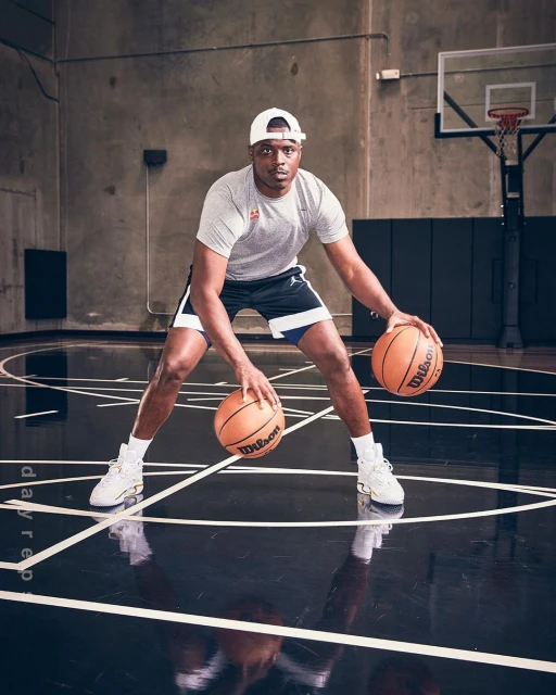 Male basketball player dribbling two basketballs on an indoor court with a concrete wall background; wearing athletic gear and a white cap.