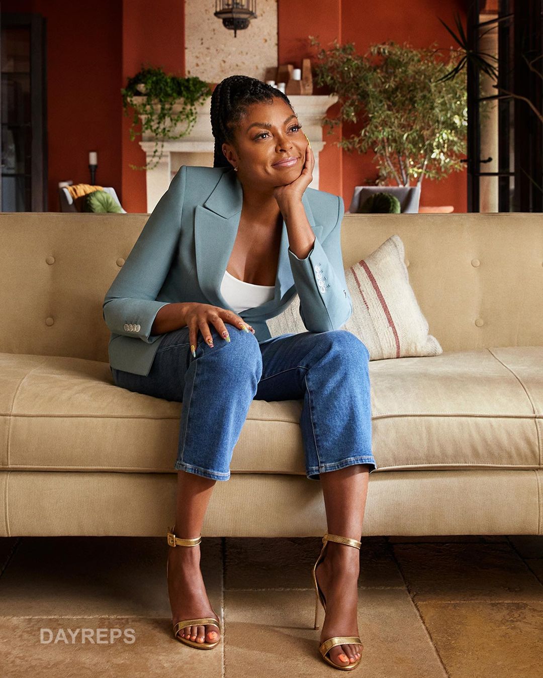 Taraji P. Henson sitting on a beige sofa in a light teal blazer and blue jeans. Her gold strappy sandals complete the look.