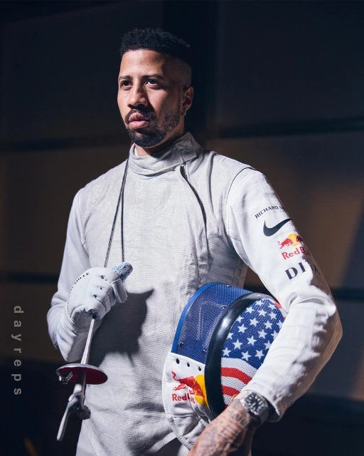 Portrait of a male fencer in uniform holding a foil and helmet with the American flag design, posing indoors with dark background.