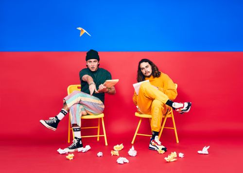 Two young adult men are seated on yellow chairs with colorful backgrounds, holding tablets surrounded by paper birds, creating a whimsical, creative scene.