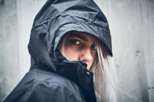 Close-up portrait of a woman with white hair wearing a black hooded coat against a textured gray wall, looking at the camera.