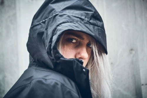 Close-up portrait of a woman with white hair wearing a black hooded coat against a textured gray wall, looking at the camera.