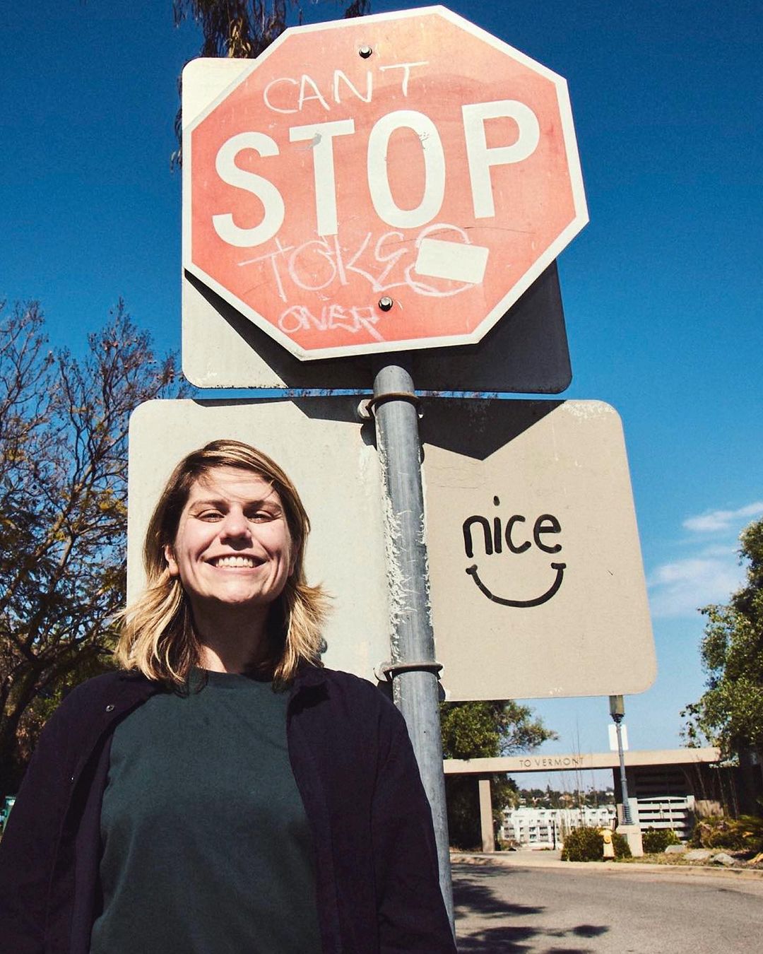 Smiling woman stands next to a stop sign defaced with graffiti and a sign that reads 'nice' on a sunny day outdoors.