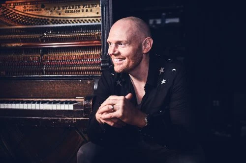 Adult male with short red hair smiles and poses next to an antique piano with a dark background in a medium shot indoors.