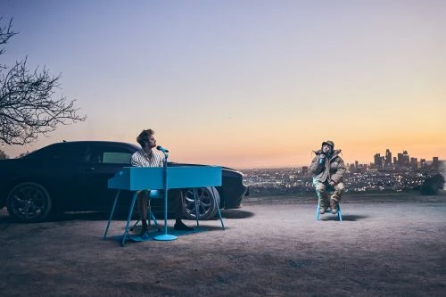 Two musicians in outdoor setting: one plays a blue keyboard next to a black car, the other sits on a stool with a city skyline background.