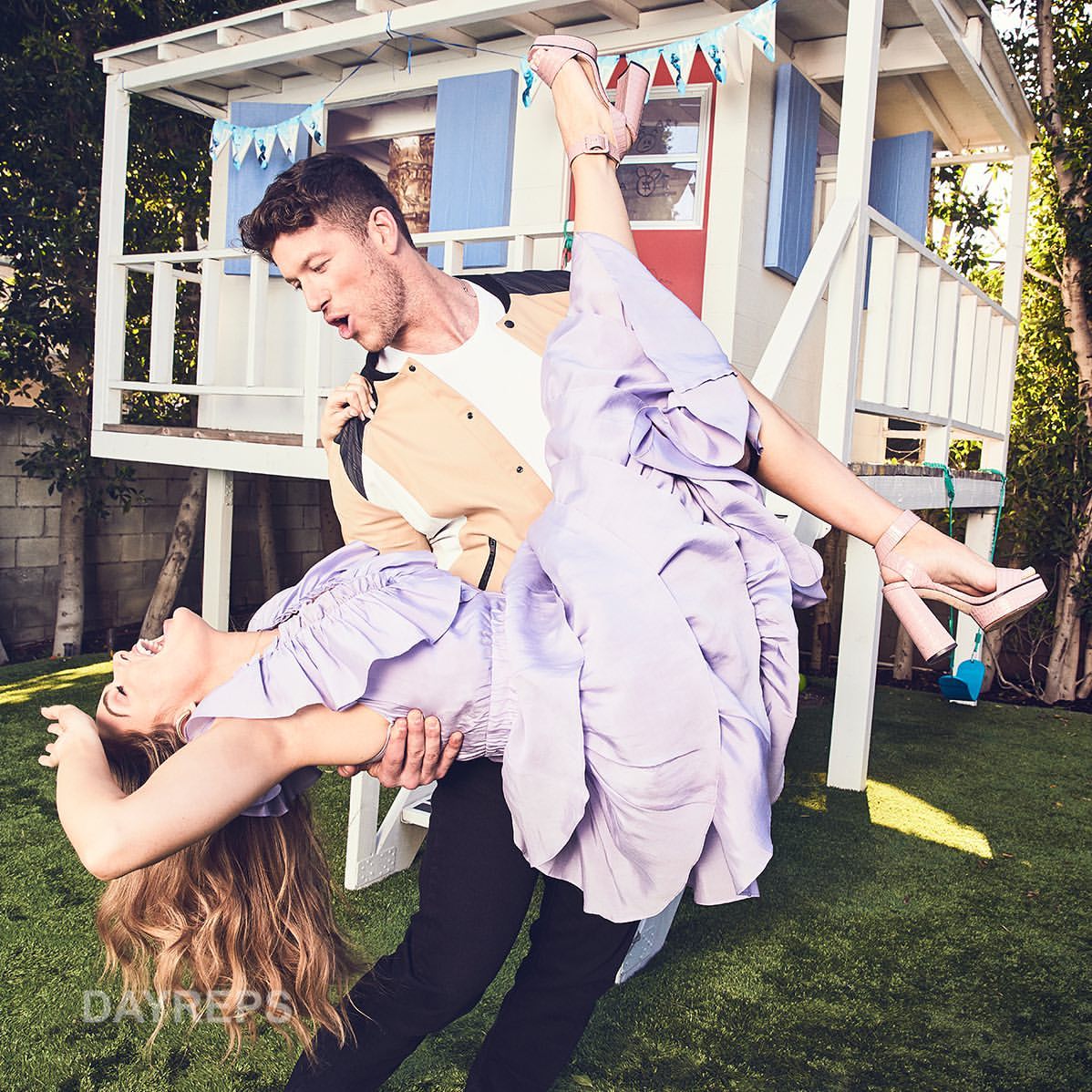 A man holds a woman upside down in a yard with a playhouse in the background; the woman is wearing a light purple dress and heels.
