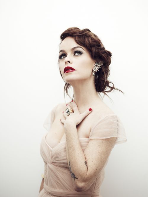 Portrait of a woman with red lipstick and her hand on her chest. She has an ornate earring, and wears a light pink gown.