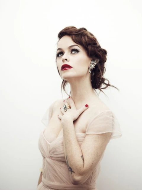 Portrait of a woman with red lipstick and her hand on her chest. She has an ornate earring, and wears a light pink gown.