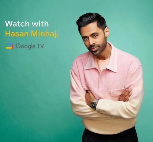 Portrait of Hasan Minhaj in a pink and cream sweater, with arms crossed, set against a teal background, promoting a Google TV show.