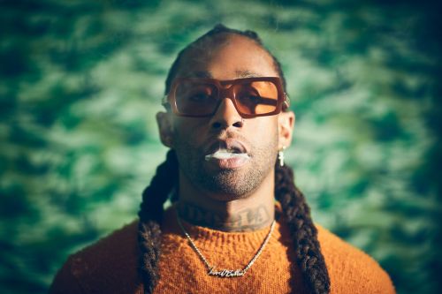 Portrait of an adult male with braided hair, brown glasses, and a mustard-colored sweater exhaling white smoke against a green backdrop.