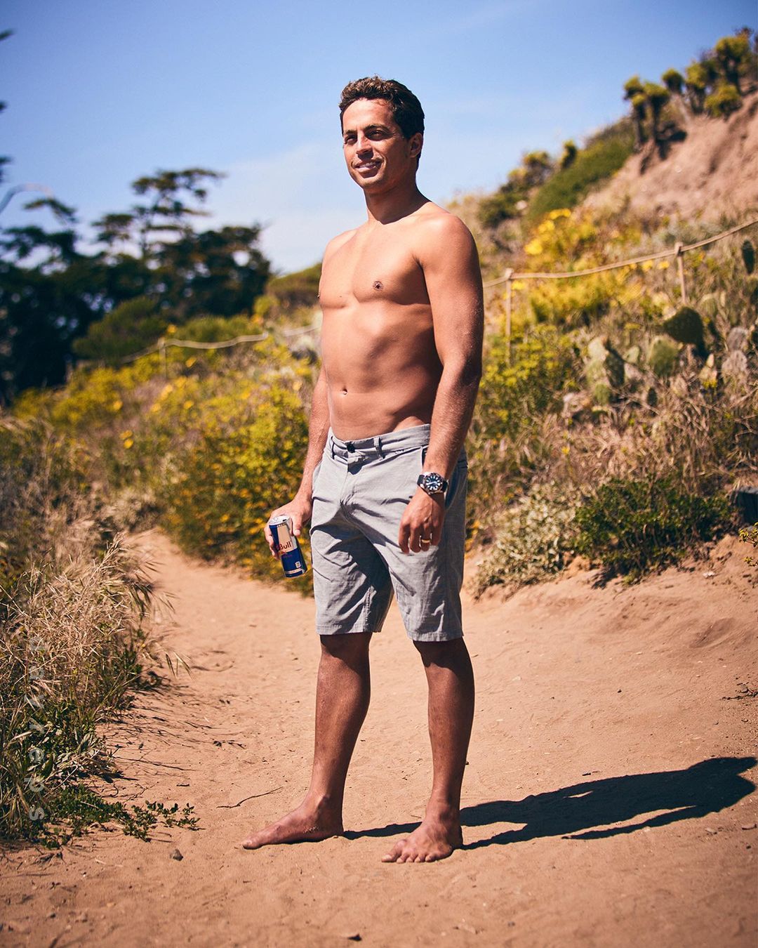 Bare-chested man stands on a sandy path near greenery, holding a can of Red Bull. The background features a blue sky and foliage.