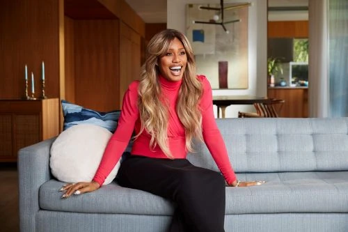Smiling adult female with long blonde hair wearing red top and black pants sitting on a couch indoors with a decorative pillow.
