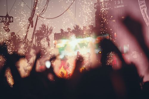 Crowd of people at an outdoor concert with stage lighting, pyrotechnics, wires and palm trees visible against a dusky sky at dusk.