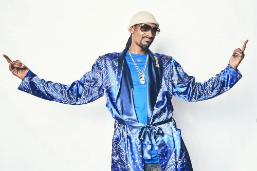 Full shot of adult male Snoop Dogg wearing a blue paisley robe, sunglasses, and a beanie while pointing both index fingers up.