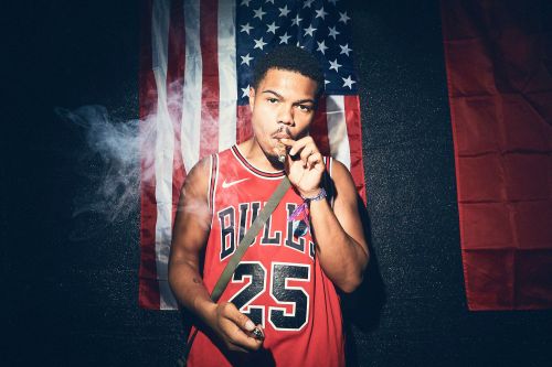 An African-American man in a red basketball jersey smokes a cigar against an American flag backdrop in an indoor setting.