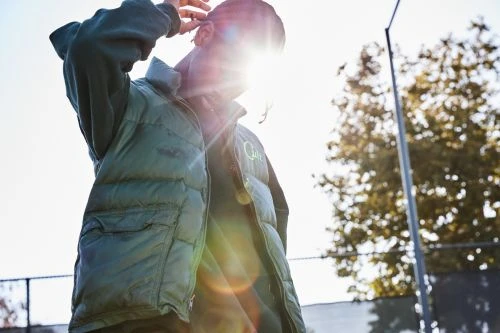 Man wearing a green puffer vest standing outdoors in Los Angeles with bright lens flare from the sun behind him.