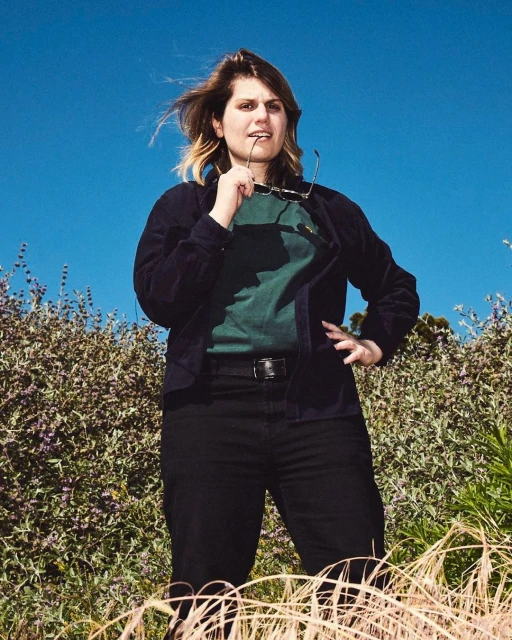 Woman poses outdoors wearing sunglasses on her mouth and a dark blue jacket against a clear blue sky with green foliage around her.