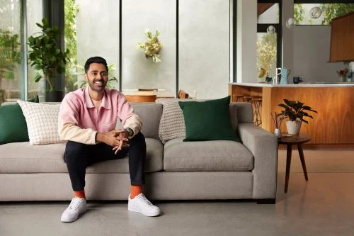Adult male sits on a neutral grey sofa in a living room setting with interior plants, side table and kitchen in background.