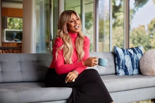 An adult female with blond wavy hair sits on a grey couch in a living room and holds a blue ceramic mug, wearing a pink top and black skirt.
