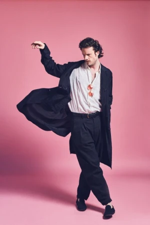 A man in a dark trench coat and white shirt dancing dynamically against a solid pink backdrop, captured by Koury Angelo.