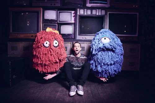 A man sits on the floor between two mop-like figures in a room filled with old televisions and media storage lockers.