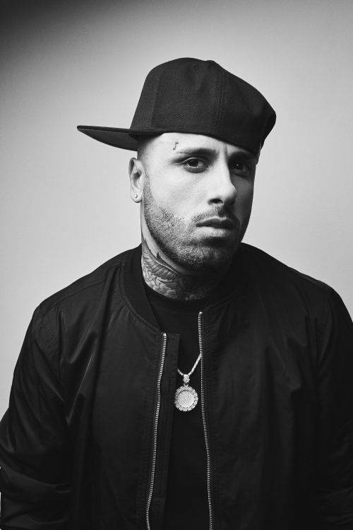Portrait of Nicky Jam wearing a black baseball cap, bomber jacket, and pendant necklace against a light background.