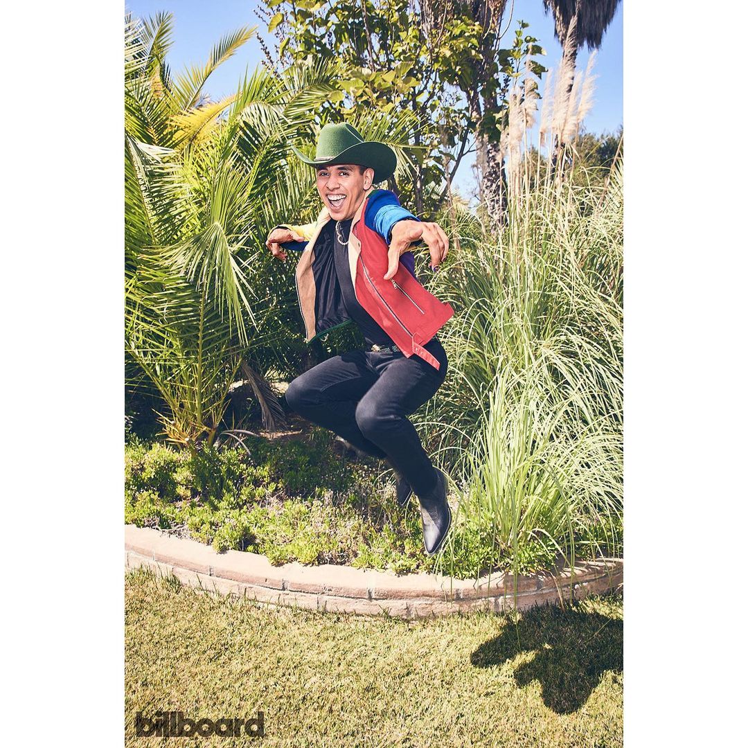 Full shot of a man in mid-air wearing a green cowboy hat, black top, and red vest in front of green plants on a sunny day.