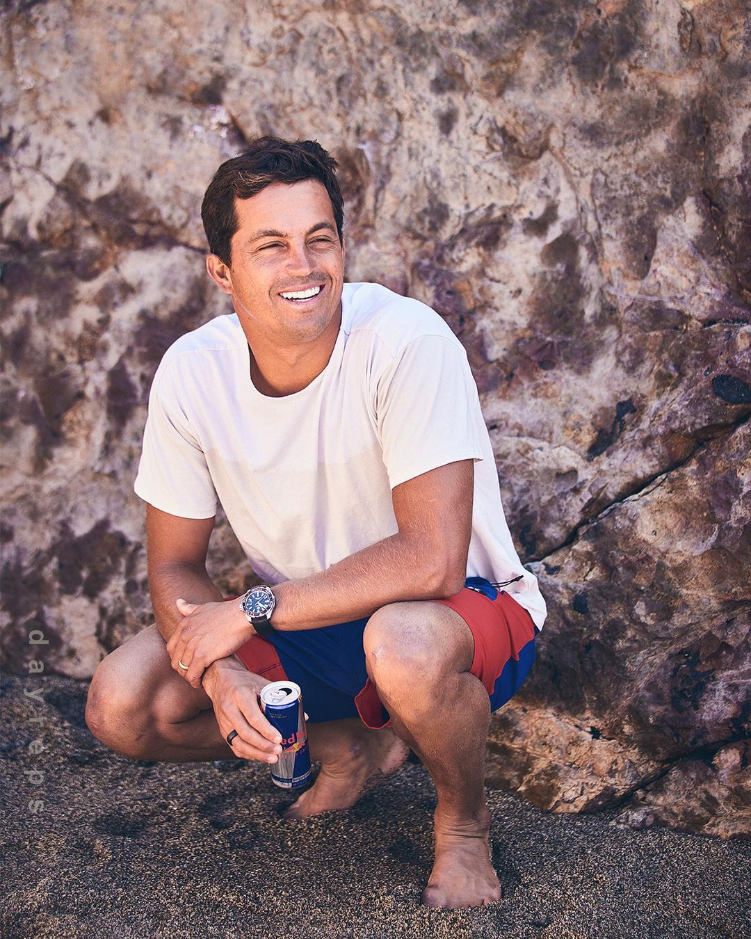 Smiling man squats on sandy beach near rock formation, holding Red Bull can, wearing white tee shirt and blue and red swim trunks.