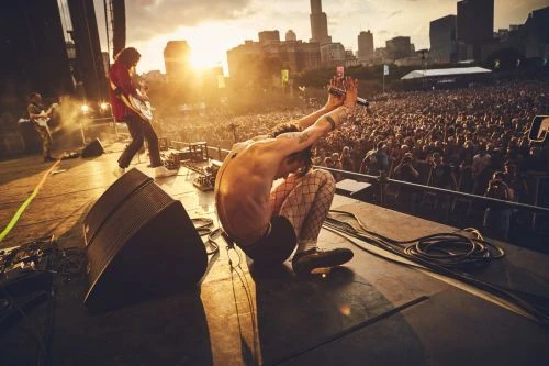 Singer performs on stage with arms raised, backed by a guitarist and a large crowd under a bright sky with city buildings visible.