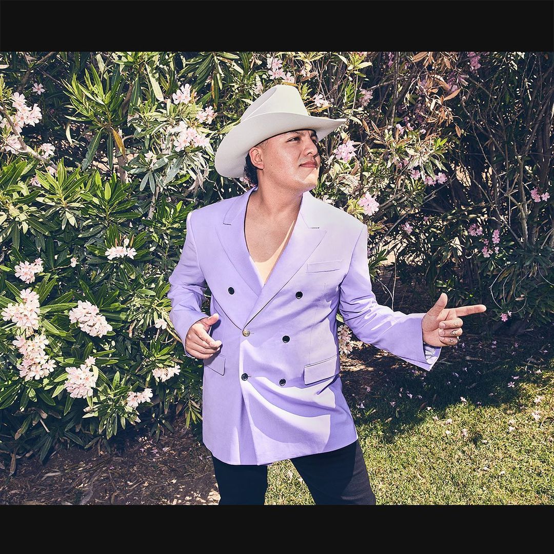 Portrait of adult male in cowboy hat and lilac suit jacket, pointing outdoors amid green foliage and white flowers in daytime lighting.