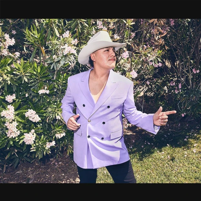 Portrait of adult male in cowboy hat and lilac suit jacket, pointing outdoors amid green foliage and white flowers in daytime lighting.