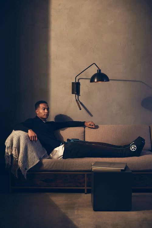 Adult male lounging on sofa in dimly lit interior. He wears dark shirt, with lower body compression-therapy device. Table lamp on wall.