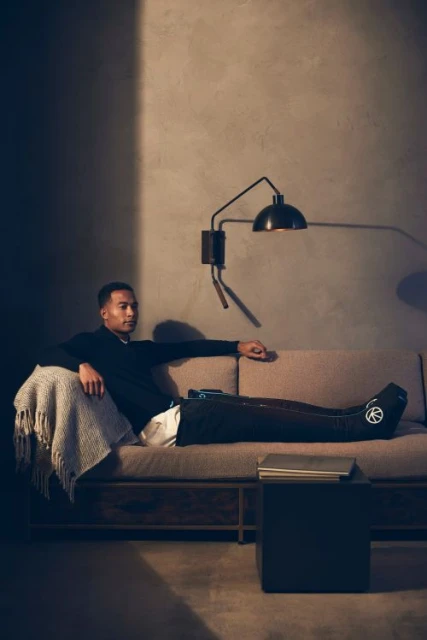 Adult male lounging on sofa in dimly lit interior. He wears dark shirt, with lower body compression-therapy device. Table lamp on wall.