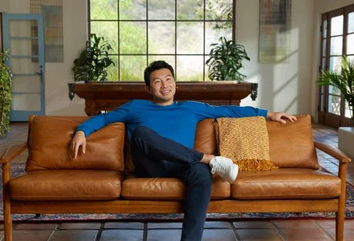 Asian man sits on a leather sofa in a modern living room, wearing a blue shirt and jeans, in a well-lit interior space with large windows.