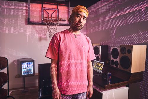 Adult male wearing a cap and pink t-shirt in a room with a basketball hoop, audio speakers, and a small refrigerator.