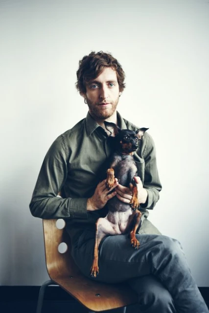 Adult male sitting on a wooden chair, holding a small black dog in a light indoor environment. Man with curly hair, wearing an olive shirt.