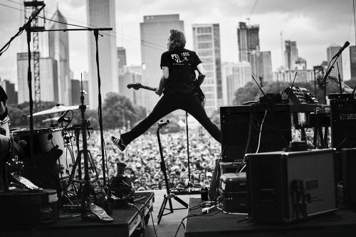 Band member mid-air jump on stage at a music festival, captured in black and white, with the crowd and city skyline in the background.