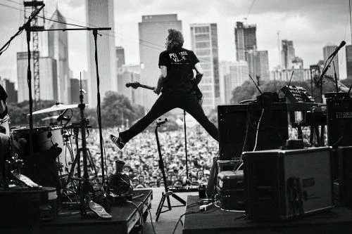 Band member mid-air jump on stage at a music festival, captured in black and white, with the crowd and city skyline in the background.