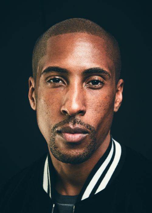 Close-up portrait of a Black man with short hair in a dark jacket, looking directly at the camera against a black background.