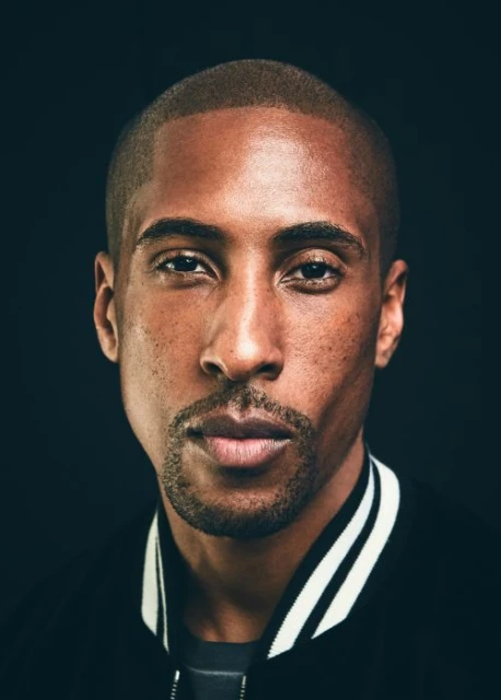 Close-up portrait of a Black man with short hair in a dark jacket, looking directly at the camera against a black background.