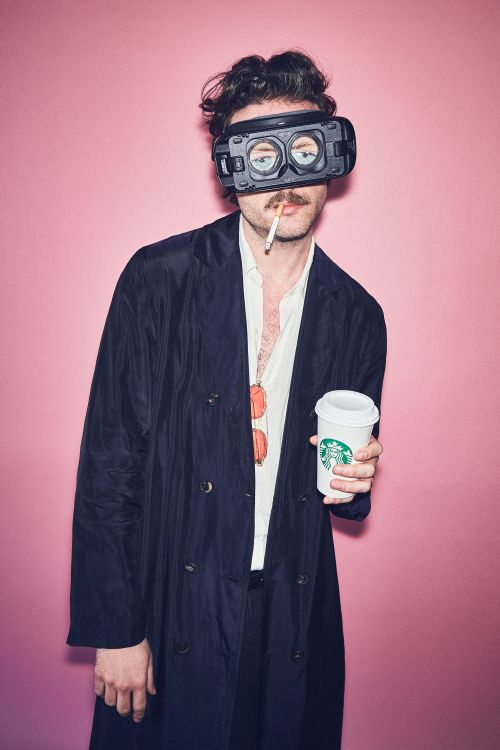 A man with a mustache and VR headset holds a lit cigarette and Starbucks coffee cup in front of a pink backdrop in a medium shot.