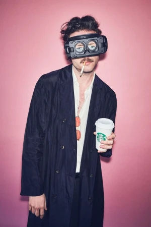 A man with a mustache and VR headset holds a lit cigarette and Starbucks coffee cup in front of a pink backdrop in a medium shot.