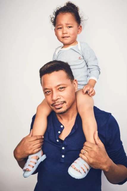 Terrance Howard carries his child on his shoulders in a studio shot. The dad wears a blue shirt, and the kid wears a gray shirt and sandals.