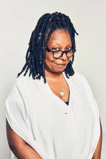 Portrait of Whoopi Goldberg with dreadlocks wearing glasses and a white blouse against a neutral backdrop in a studio setting.