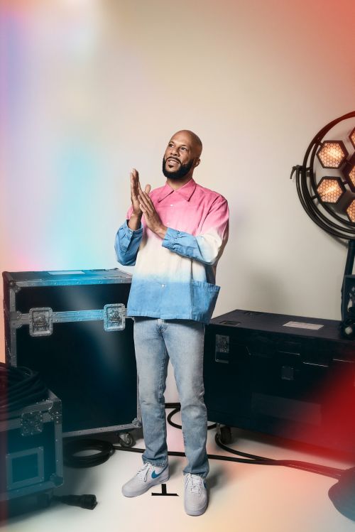 Portrait of a bald adult male clapping his hands, wearing a light pink and blue shirt and jeans, with stage equipment behind him.