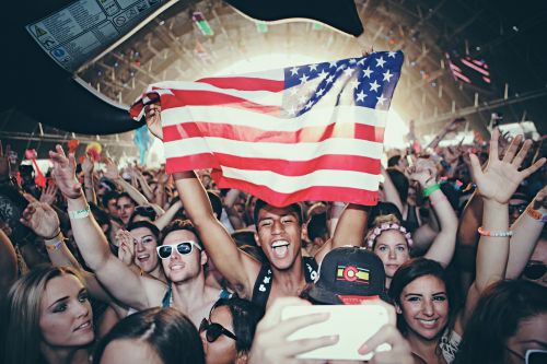 Crowd of people cheering at an outdoor music festival, with a man holding an American flag aloft in the center of the frame.