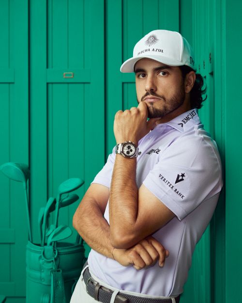 Portrait of a male golfer in a baseball cap leaning against a green wall next to golf clubs, wearing a lavender shirt and wristwatch.